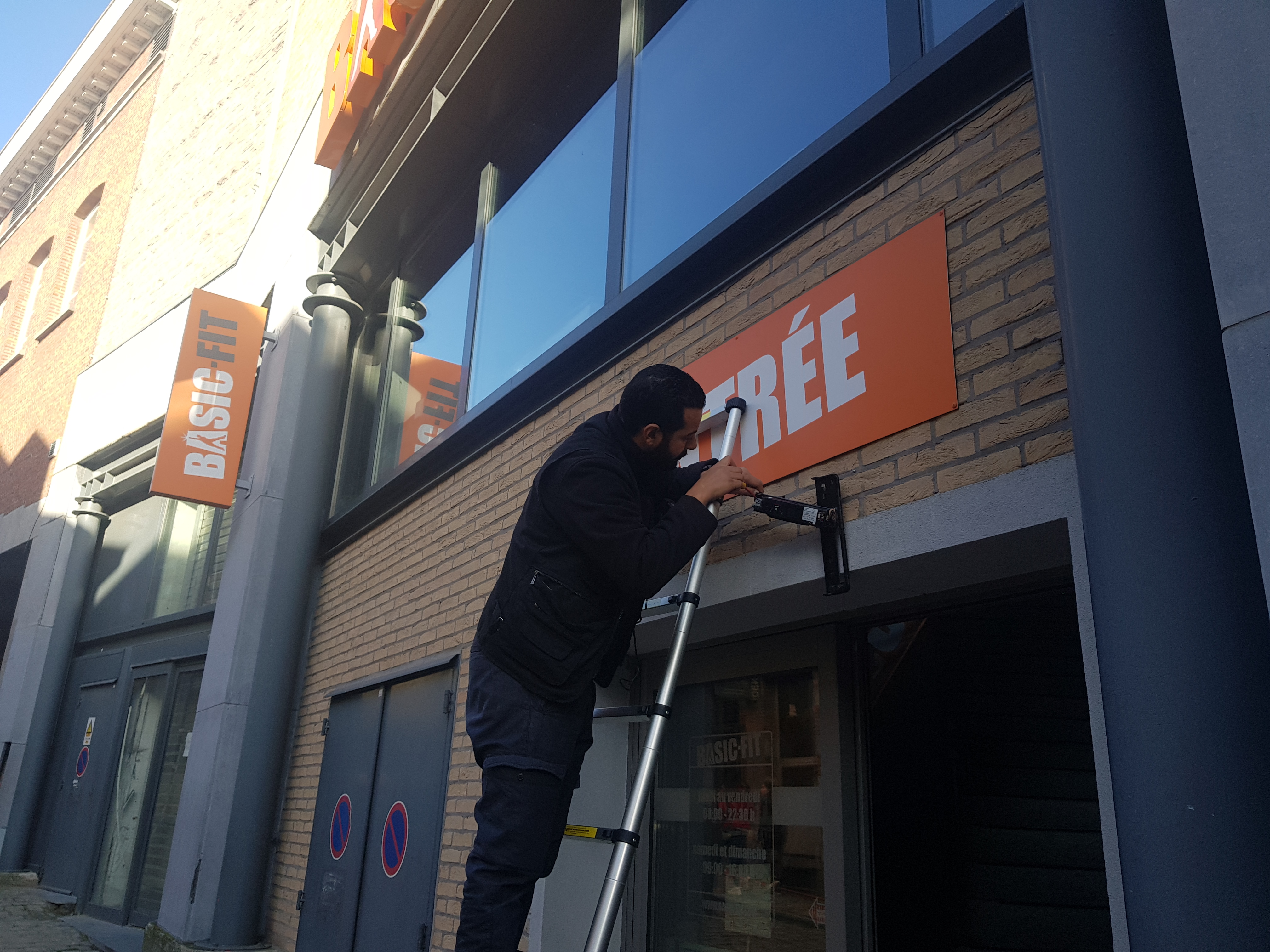 Technicien ELECOMM sur échelle remontant le capteur de mouvement en façade du Basic-Fit Mons